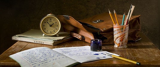 old fashioned analog clock, notebooks, pencils, inkwell, and leather briefcase arranged on worn wooden table